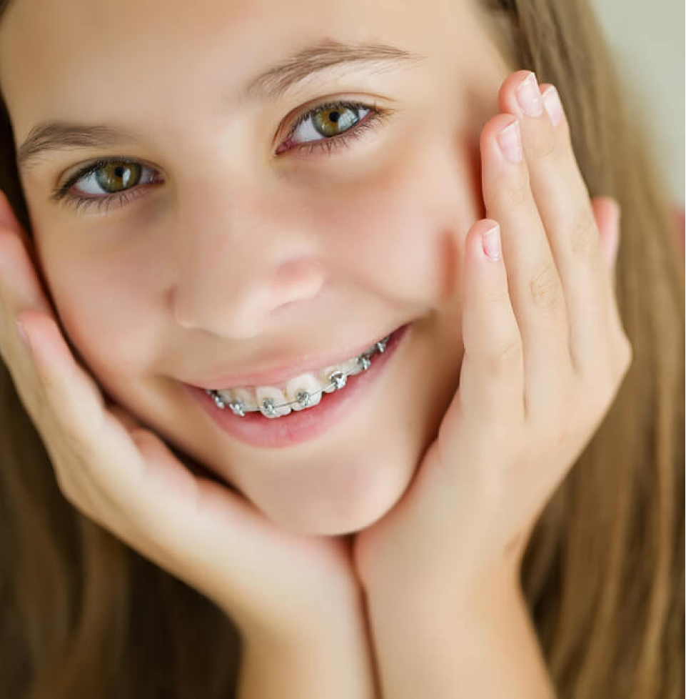 Young girl with colorful braces smiling at camera, hands to face, indoor portrait.
