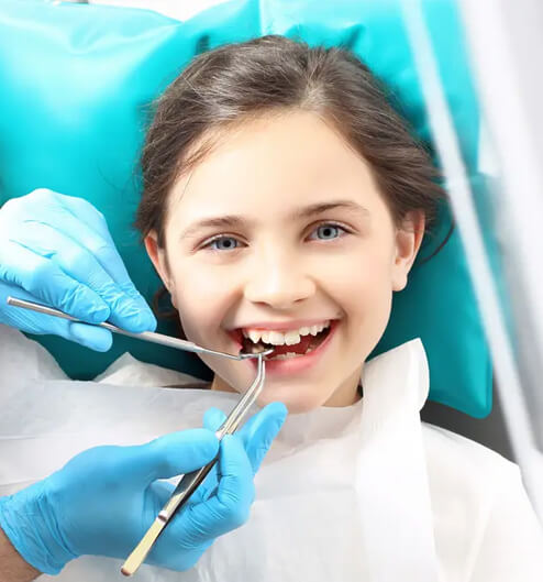 Young girl smiling at dental checkup with dentist holding tools and wearing blue gloves.