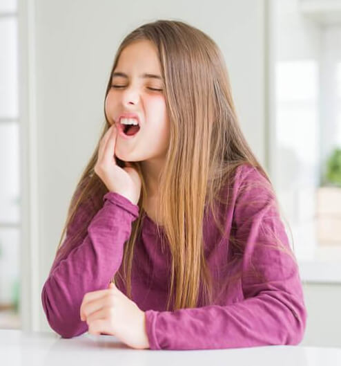 Young girl in purple shirt smiling with hand to face in bright dental office setting.