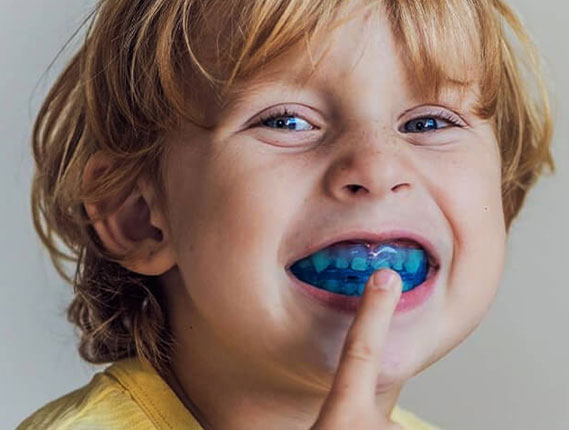 Young child with curly red hair smiling while brushing teeth with blue toothbrush.