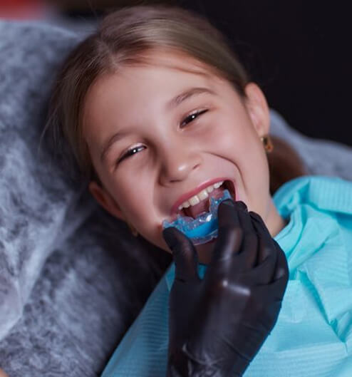 Young girl in blue jacket eating blue popsicle, wearing black gloves, smiling indoors.