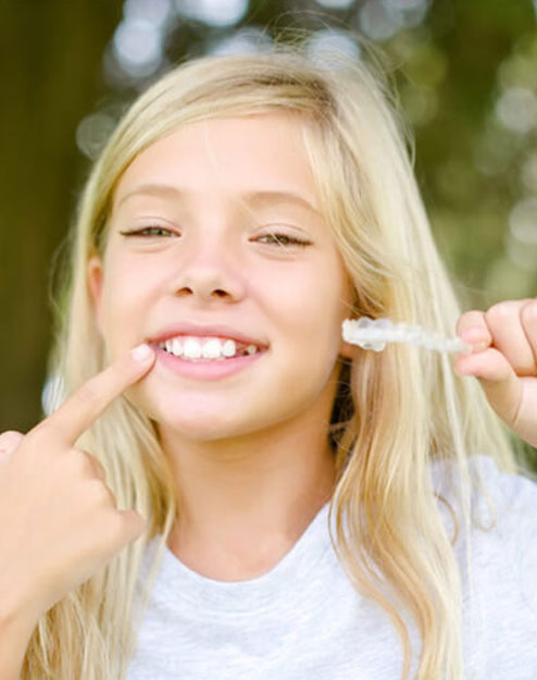 Young girl smiling brightly while pointing to her teeth outdoors in natural daylight.