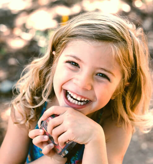 Happy young girl smiling brightly, holding blue toothbrush outdoors in natural setting.