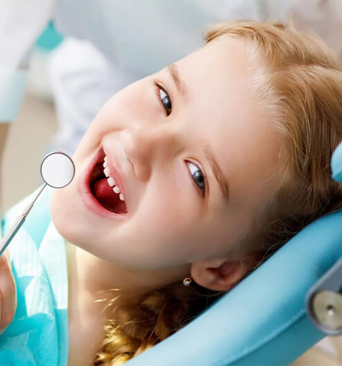 Young child smiling in dental chair with mirror during pediatric dental examination.