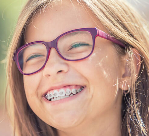 Young girl wearing purple glasses smiling, displaying braces in dental office setting.