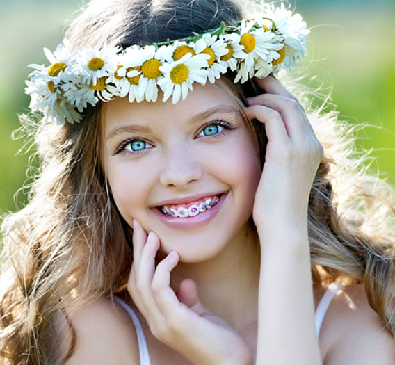 Young girl wearing flower crown smiling outdoors in natural sunlight displaying healthy teeth.