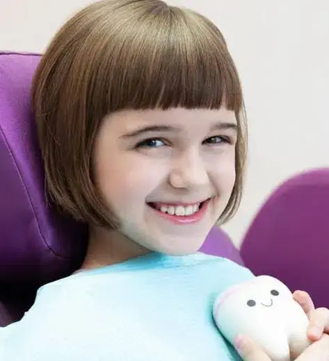 Happy child in light blue shirt holding stuffed animal in dental office chair.
