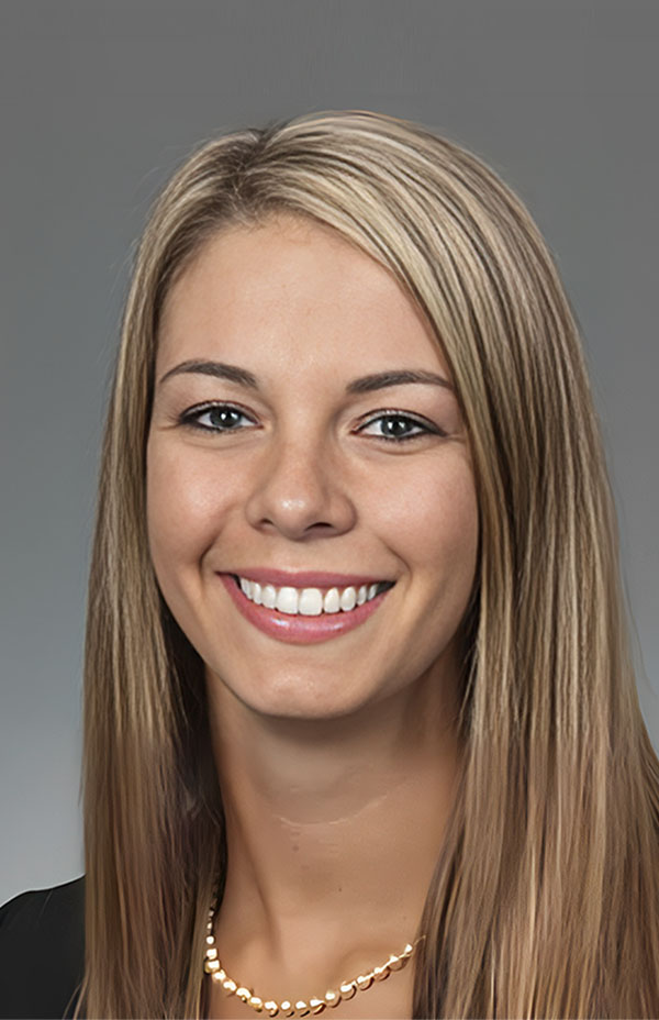 Professional headshot of smiling woman with long blonde hair wearing pearl necklace against gray background.