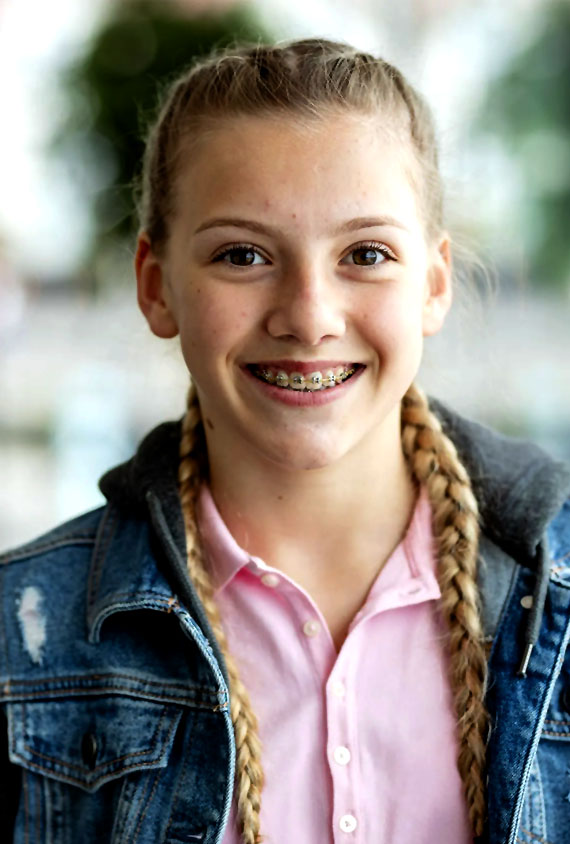 Young girl with braided hair smiling at camera wearing denim jacket and pink shirt.