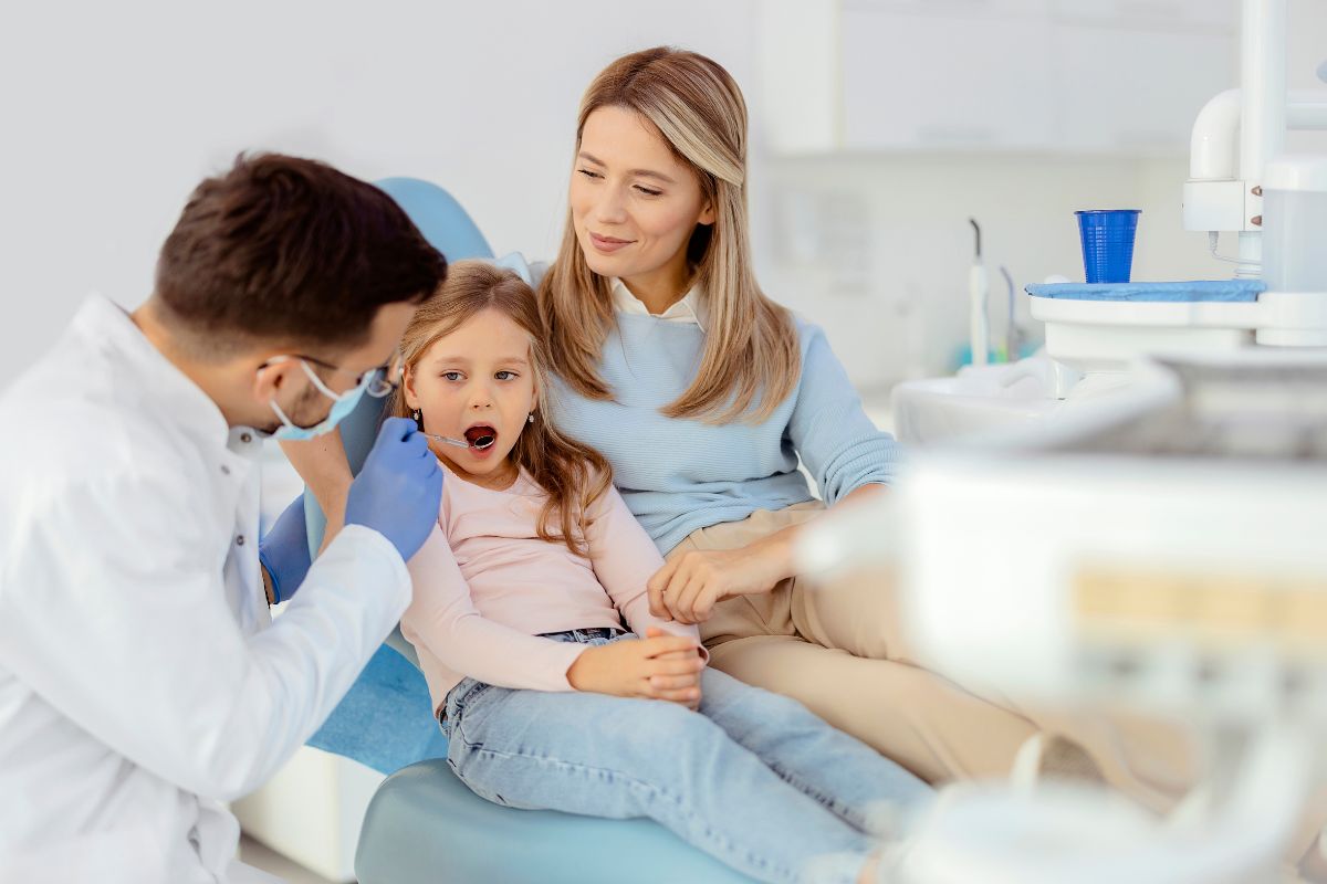 Pediatric dentist examining young child's teeth while parent watches in modern dental office.